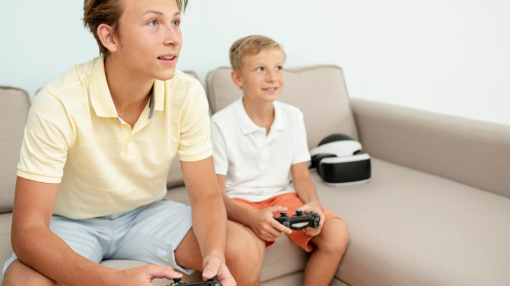 Dos hermanos, uno más mayor que otro, juegan al mismo videojuego sentados en el sofá