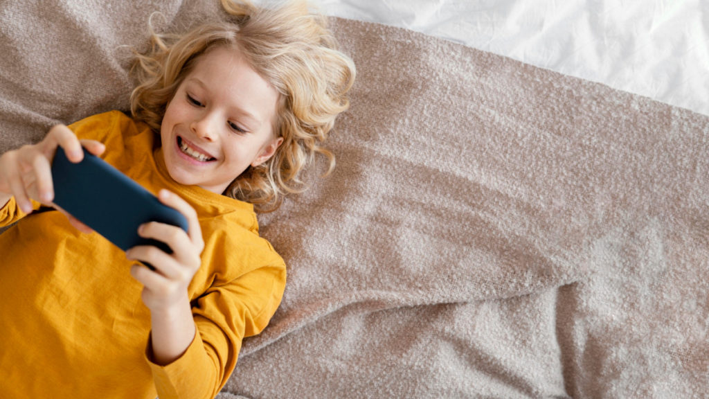 Un niño juega a videojuegos con el teléfono móvil tumbado en la cama
