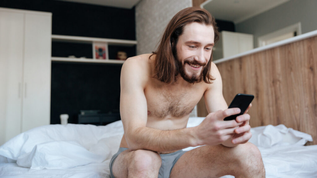 Un hombre contento y semidesnudo mira el teléfono desde la cama.