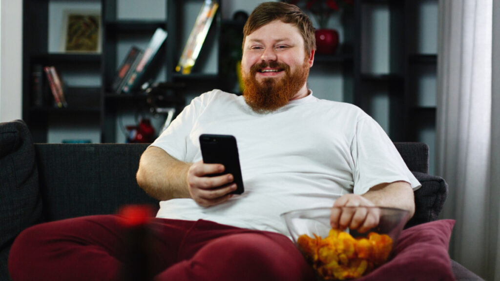 Un hombre con sobrepeso come comida basura sentado en el sillón, con un riesgo mayor de grasa visceral