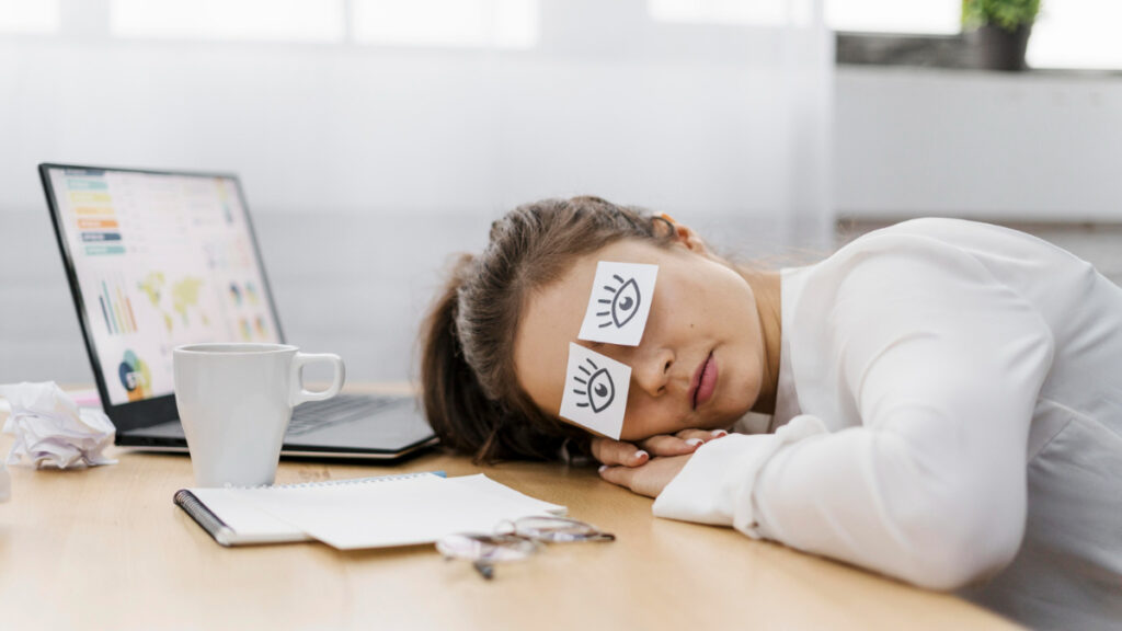 Una mujer dormida en el puesto de trabajo con pegatinas de ojos abiertos sobre los párpados