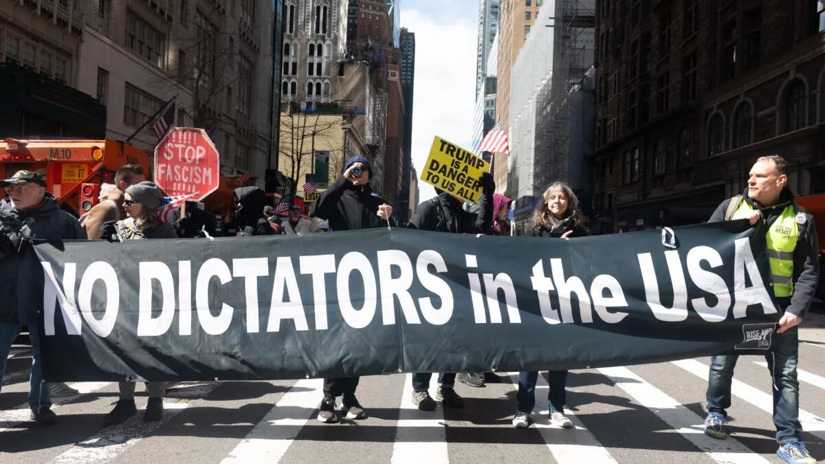 Un grupo de personas andan por la calle en Nueva York, blandiendo pancartas. En primer plano, una pancarta negra con texto blanco que reza "Nada de dictadores en EEUU"
