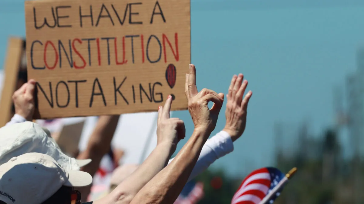 Una serie de manos con banderas estadounidenses desenfocadas. Se ve una pancarta que reza "Tenemos una constitución, no un rey"