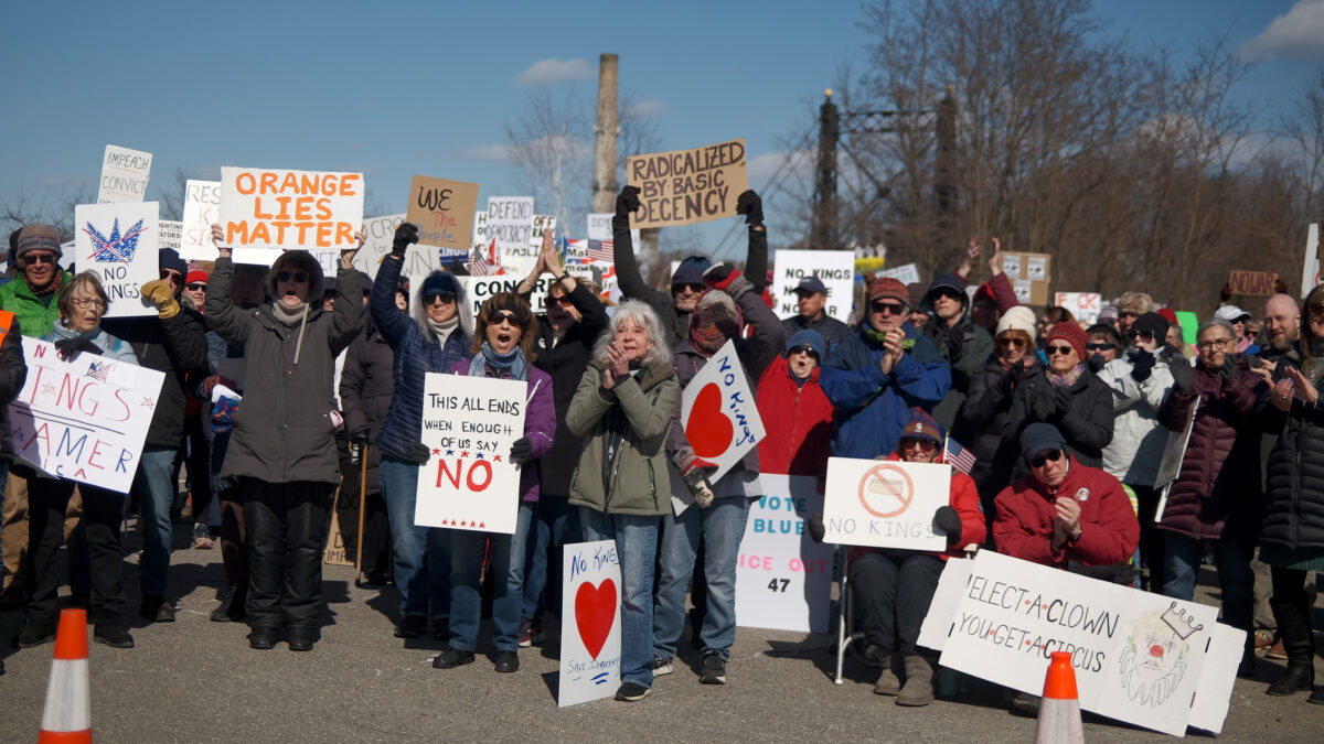 Un grupo de personas mira a cámara y blande pancartas, todas contentas, manifestándose contra el presidente estadounidense