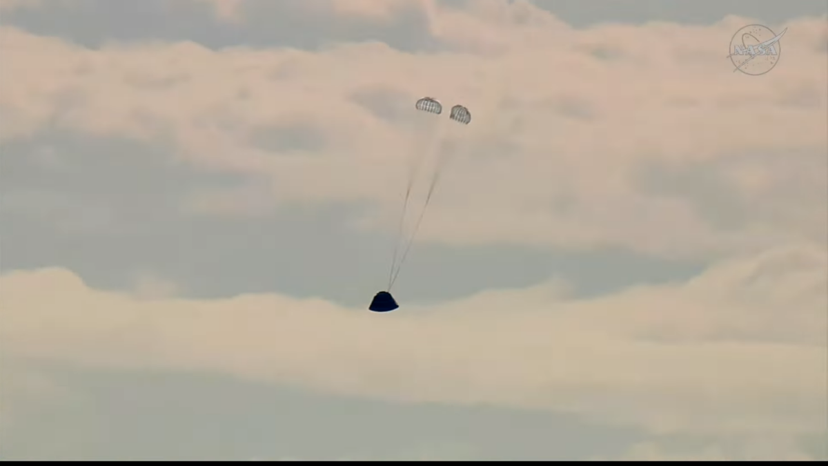 Una cápsula cónica delante de nubes, tiene dos pequeños paracaídas semiesféricos unos metros por encima, empezando a frenarla