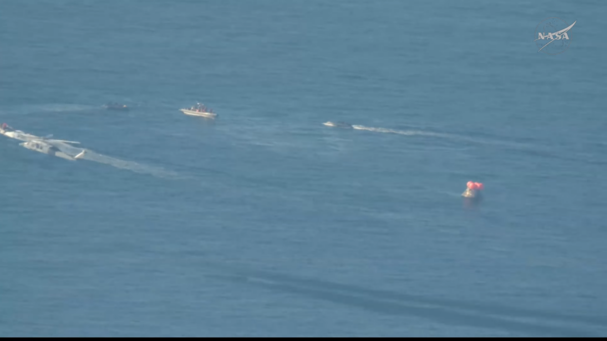 Una cápsula cónica flota en el mar. A su alrededor, varios barcos rápidos y un helicóptero